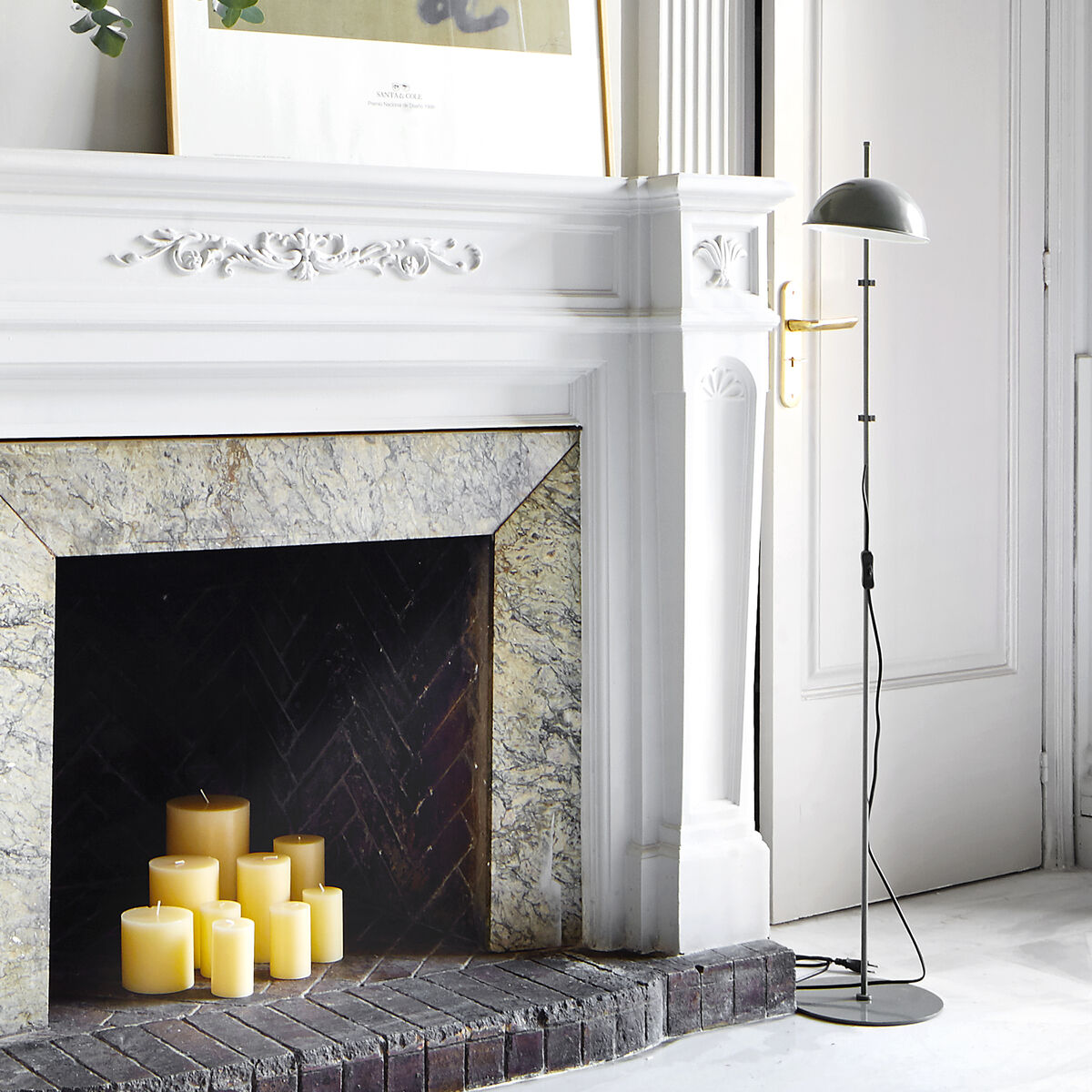 Eine moderne Stehleuchte »Funiculi« in einem minimalistisch eingerichteten Wohnzimmer mit weißem Kamin, daneben eine Tür und darüber ein gerahmtes Kunstwerk. Kerzen sind im Kamin angeordnet.