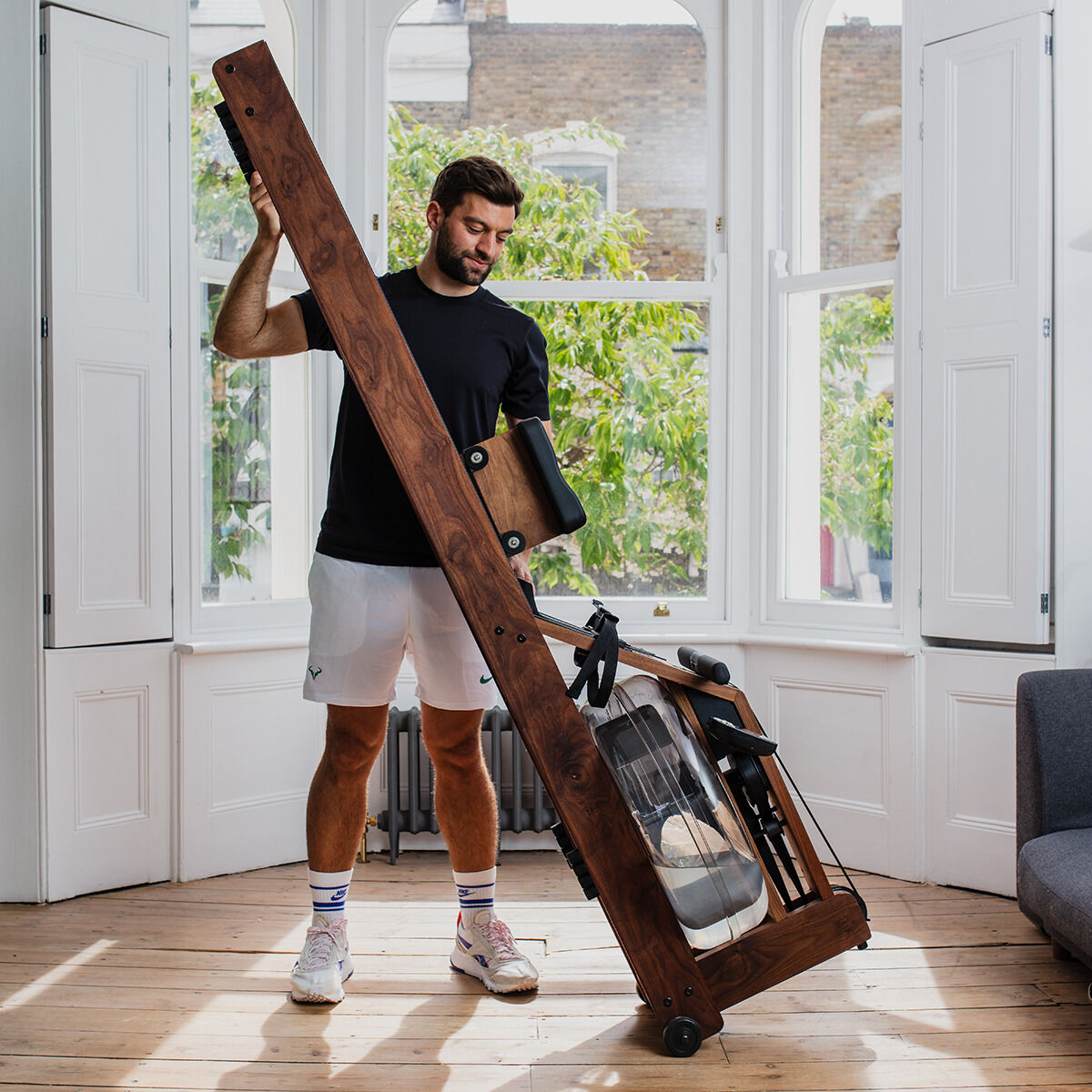 Ein Mann steht in einem hellen Raum mit einem Waterrower-Rudergerät aus Eichenholz. Der Boden ist aus Holz, und durch die großen Fenster ist grünes Laub zu sehen. Der Mann trägt Sportkleidung.