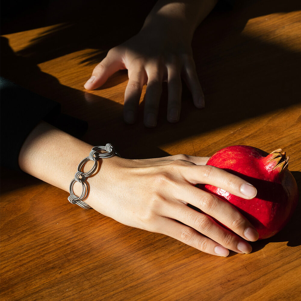 Eine Hand mit einem silbernen Armband »OMA« hält einen roten Apfel auf einem Holztisch.