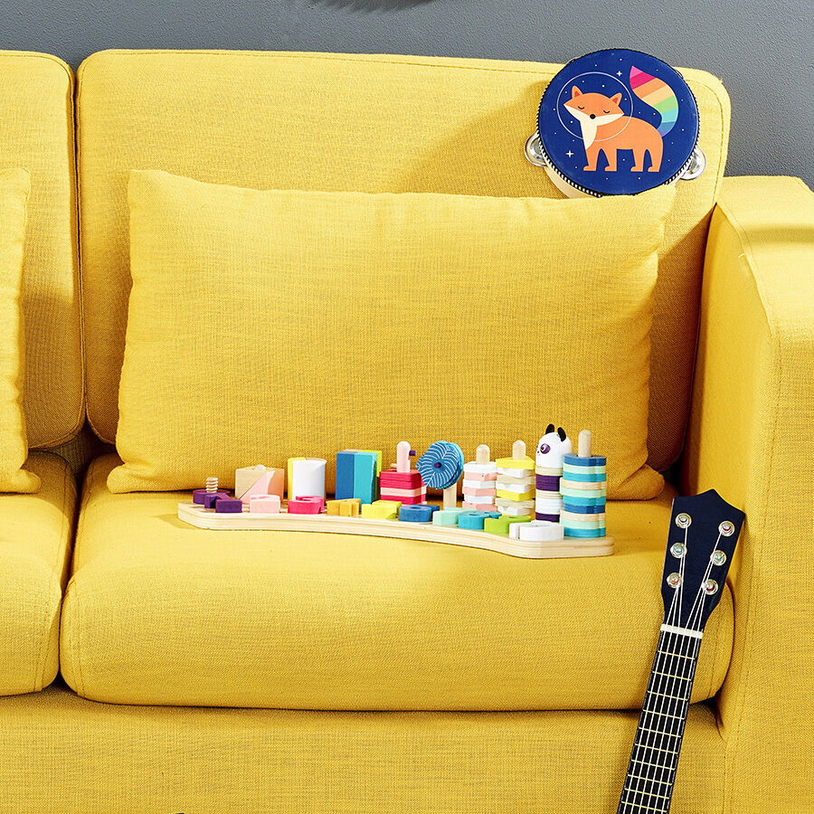 Ein Zahlenlernspiel aus Holz präsentiert sich auf einem gelben Sofa. Das Spielzeug enthält bunte, stapelbare Elemente in verschiedenen Farben und Formen. Im Hintergrund liegt ein rundes Kissen mit einem Fuchs-Motiv darauf. Rechts unten im Bild ist ein Teil einer Ukulele zu sehen.