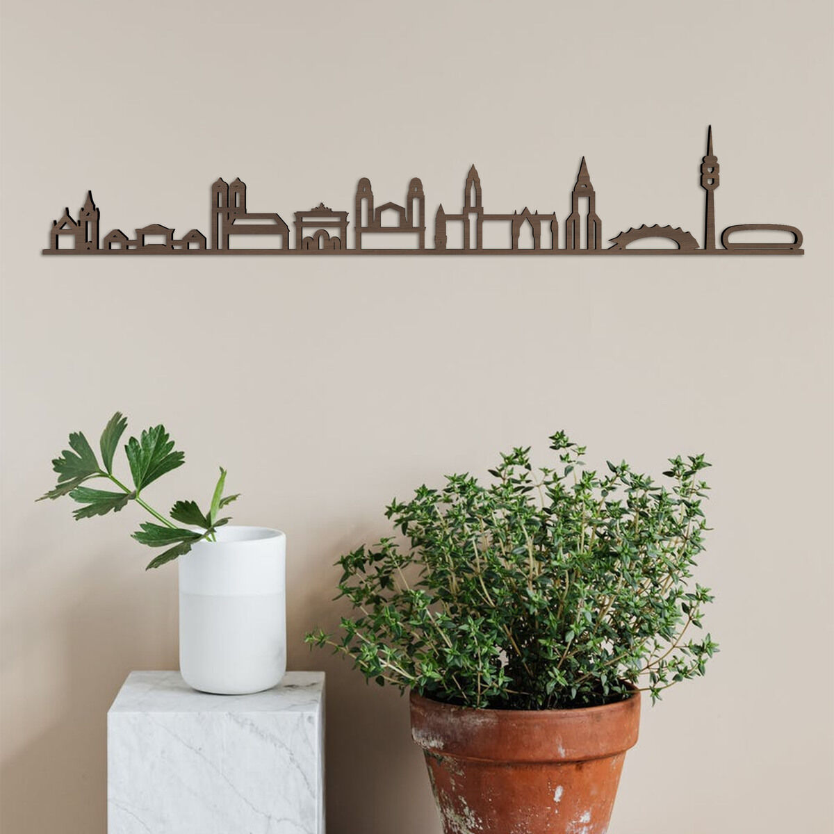 Skyline aus Holz von München, an einer beigefarbenen Wand montiert. Darunter stehen eine weiße Vase mit grünen Pflanzen auf einem weißen Sockel und ein großer Blumentopf mit einer größeren Grünpflanze.
