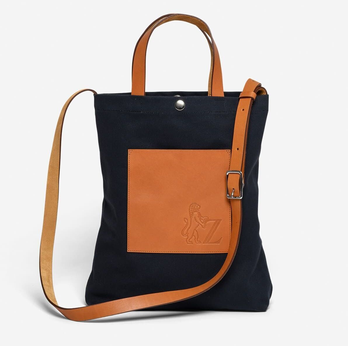 Schwarze Einkaufstasche aus Stoff mit braunen Ledergriffen und einer braunen aufgesetzten Leder-Tasche mit einem geprägten Löwen-Logo und einem verstellbaren Schulterriemen.
