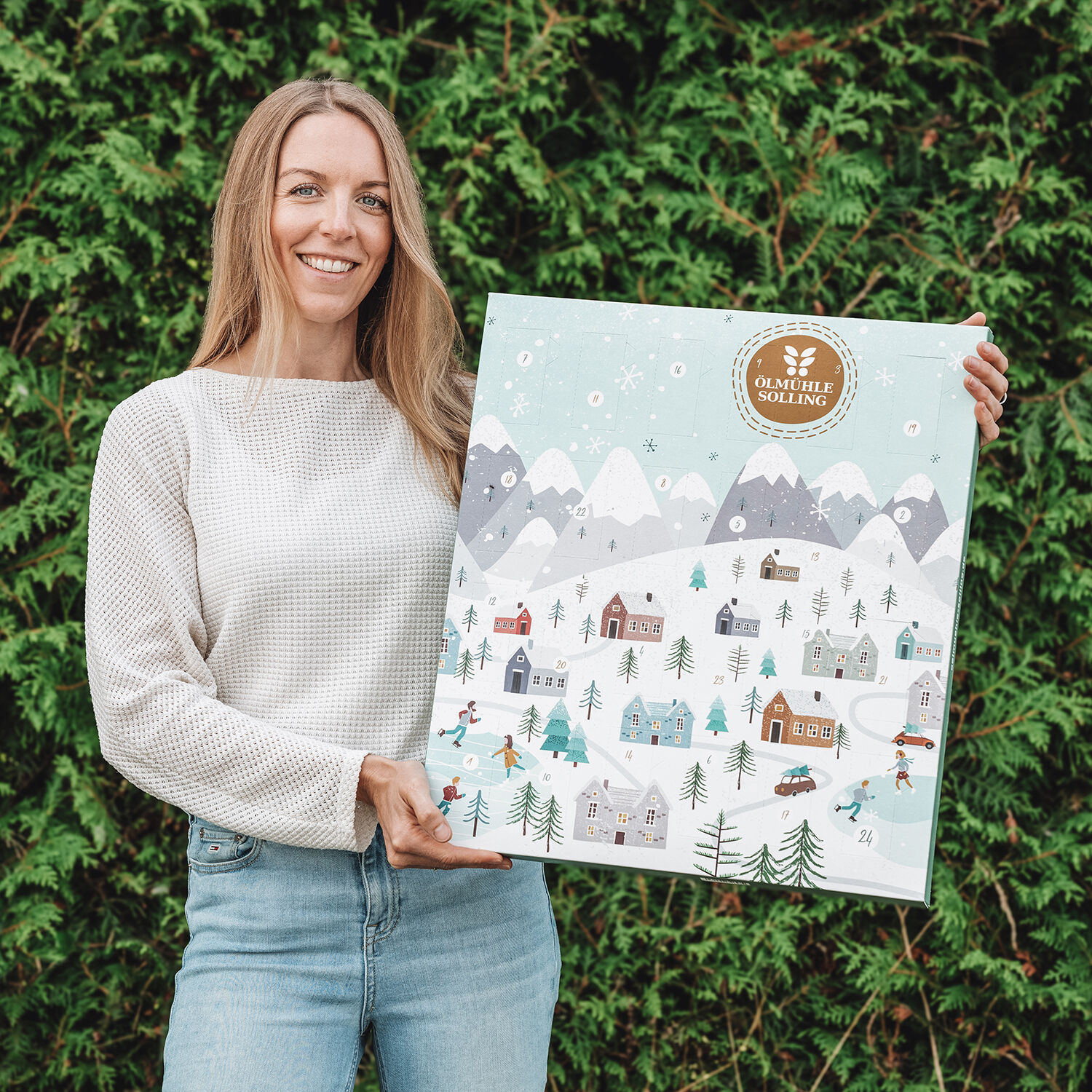 Eine lächelnde Frau hält einen großen Adventskalender mit winterlicher Schlittschuhlauf-Szenerie, verschneiten Häusern und Bergen vor einem grünen Hintergrund in den Händen.