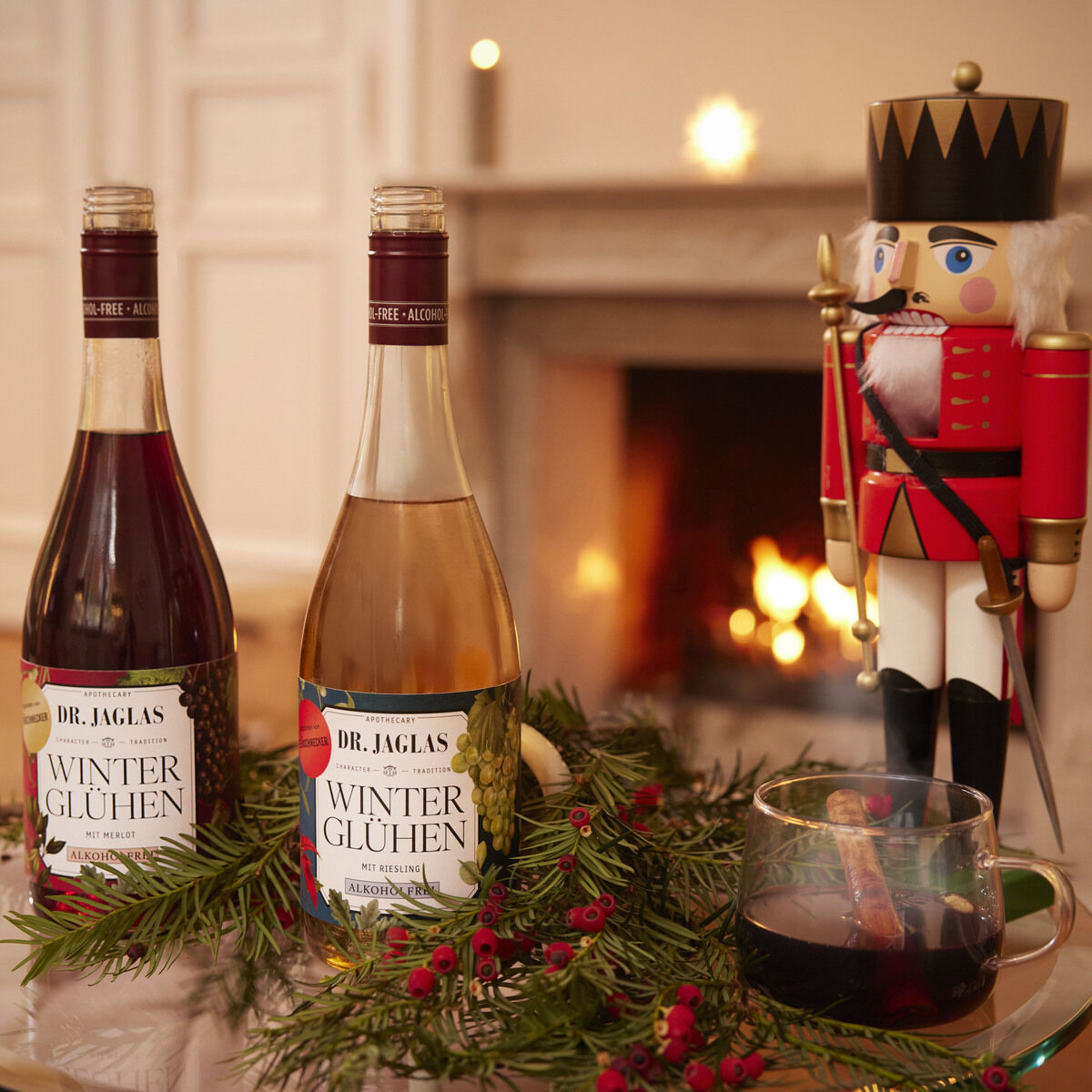 Auf einem Glastisch vor einem Kamin mit Feuer stehen zwei Flaschen DR. JAGLAS Winterglühen Rot +Weiß alkoholfreier Winzerglühwein, dekoriert mit Tannenzweigen und roten Beeren. Rechts davon ist eine Holz-Nussknackerfigur, daneben ein Glas mit Glühwein. Die Szene wirkt festlich und gemütlich.