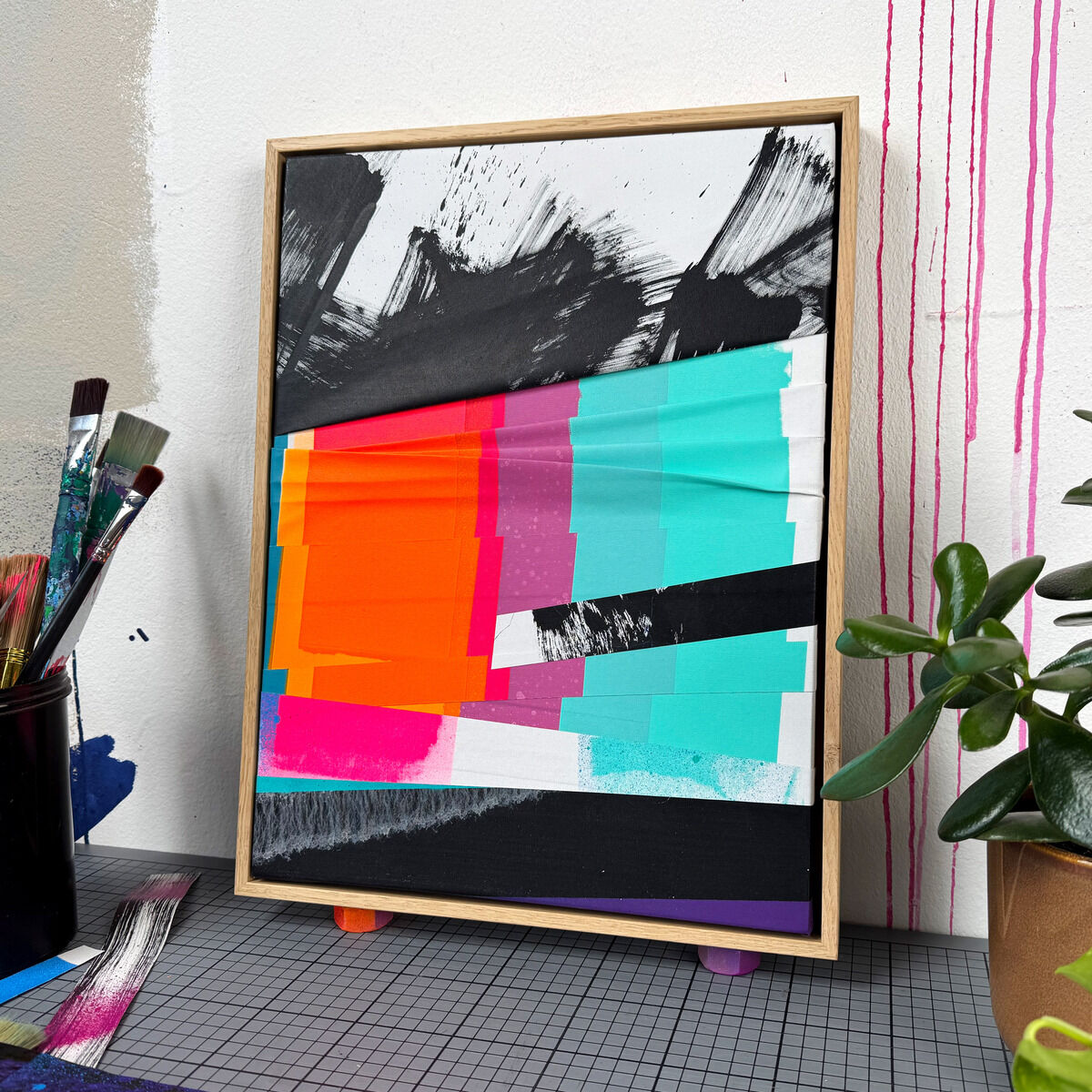 Das Unikat »Gratitude - No.3« von Daniel Lisson zeigt eine abstrakte Collage mit leuchtenden türkis-, pink- und orangefarbenen Flächen, ergänzt von dynamischen schwarzen und weißen Elementen. Gerahmt in geölter Eiche, steht es auf einem Arbeitstisch vor einem weißen Hintergrund.