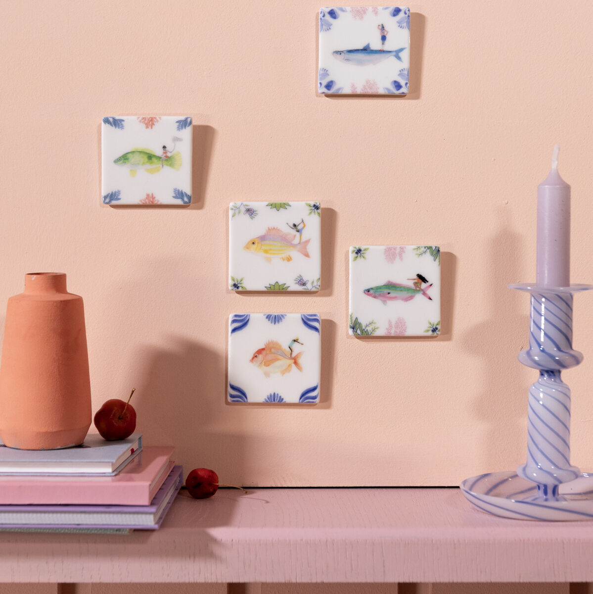 Keramikfliesen-Set »Blick nach vorn« mit floralen und tierischen Mustern an einer hellrosa Wand, begleitet von einer lila Kerze und einem orangen Dekorationsgegenstand.