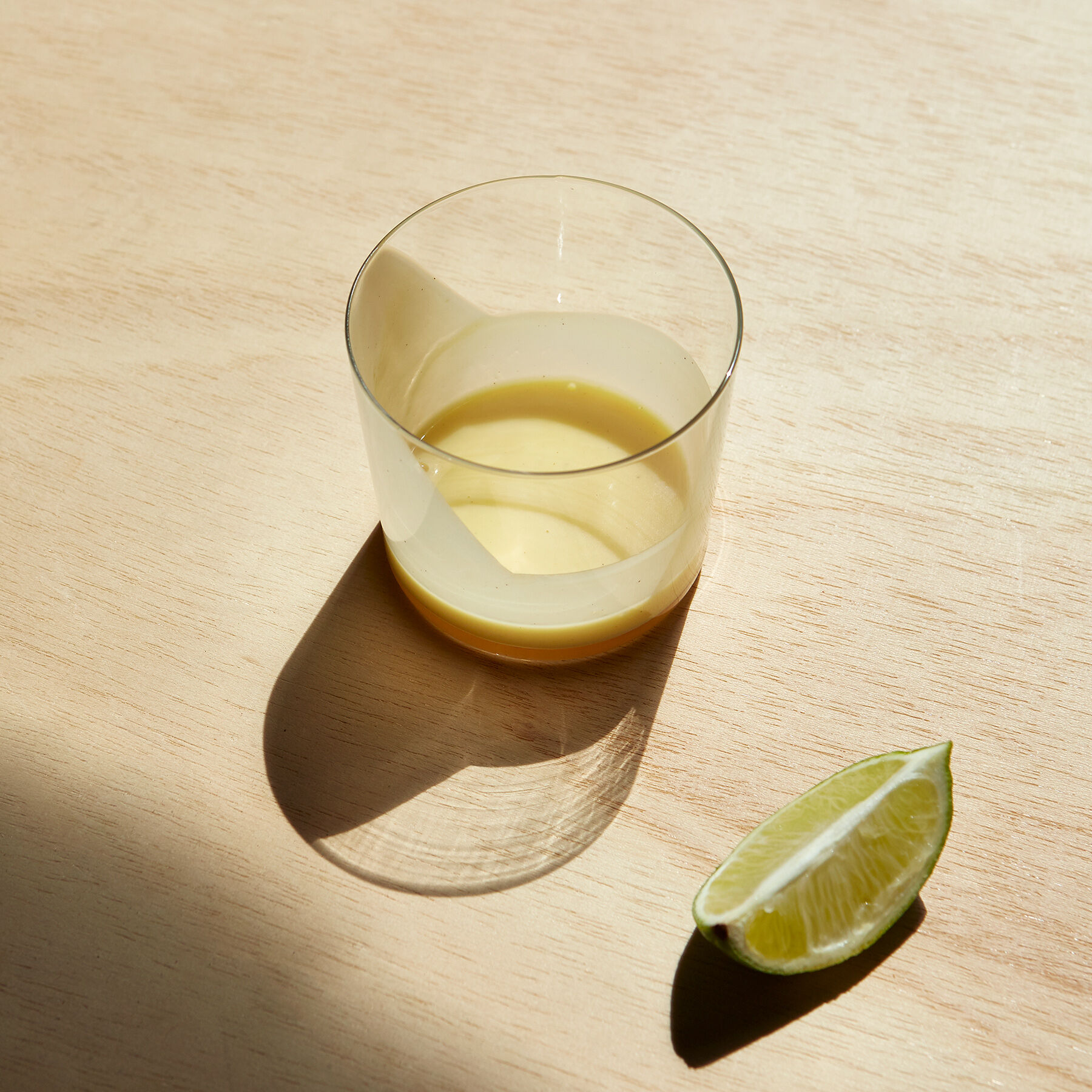 Ein Glas mit Eierlikör steht auf einer Oberfläche aus hellem Holz, daneben liegt ein Stück Limette. Die Szene ist von Sonnenlicht erhellt.