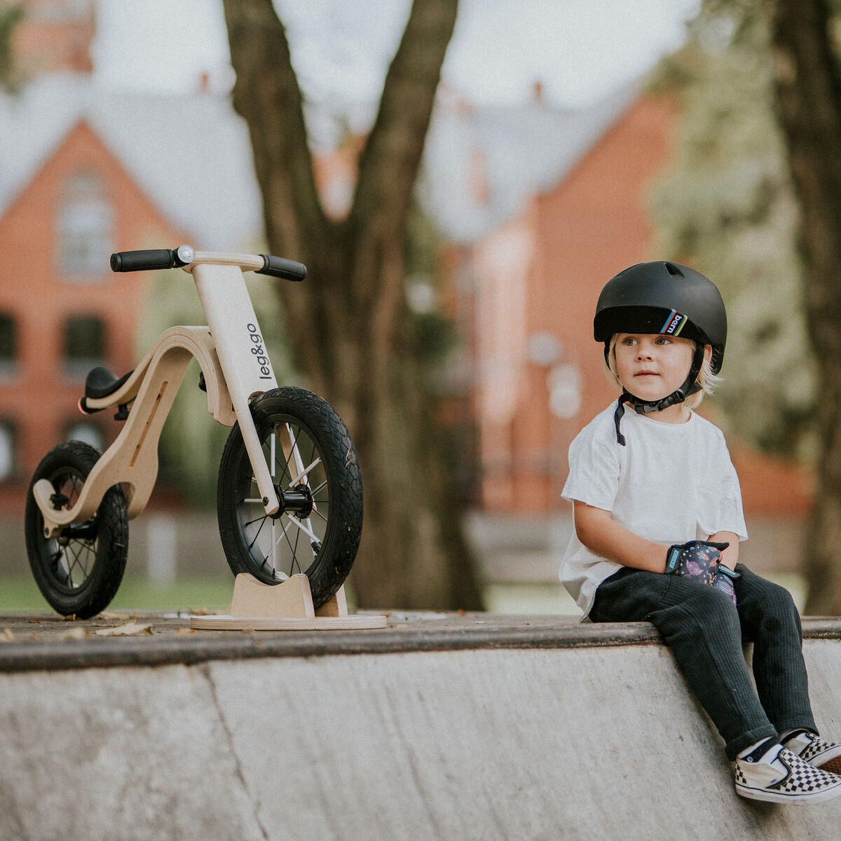 Ein Kind mit Helm sitzt auf einer Rampe im Park neben einem Balance Bike 3in1 aus Holz. Im Hintergrund sind Bäume und Gebäude zu sehen. Das Laufrad steht aufrecht und ist auf den urbanen Gebrauch im Freien ausgelegt.
