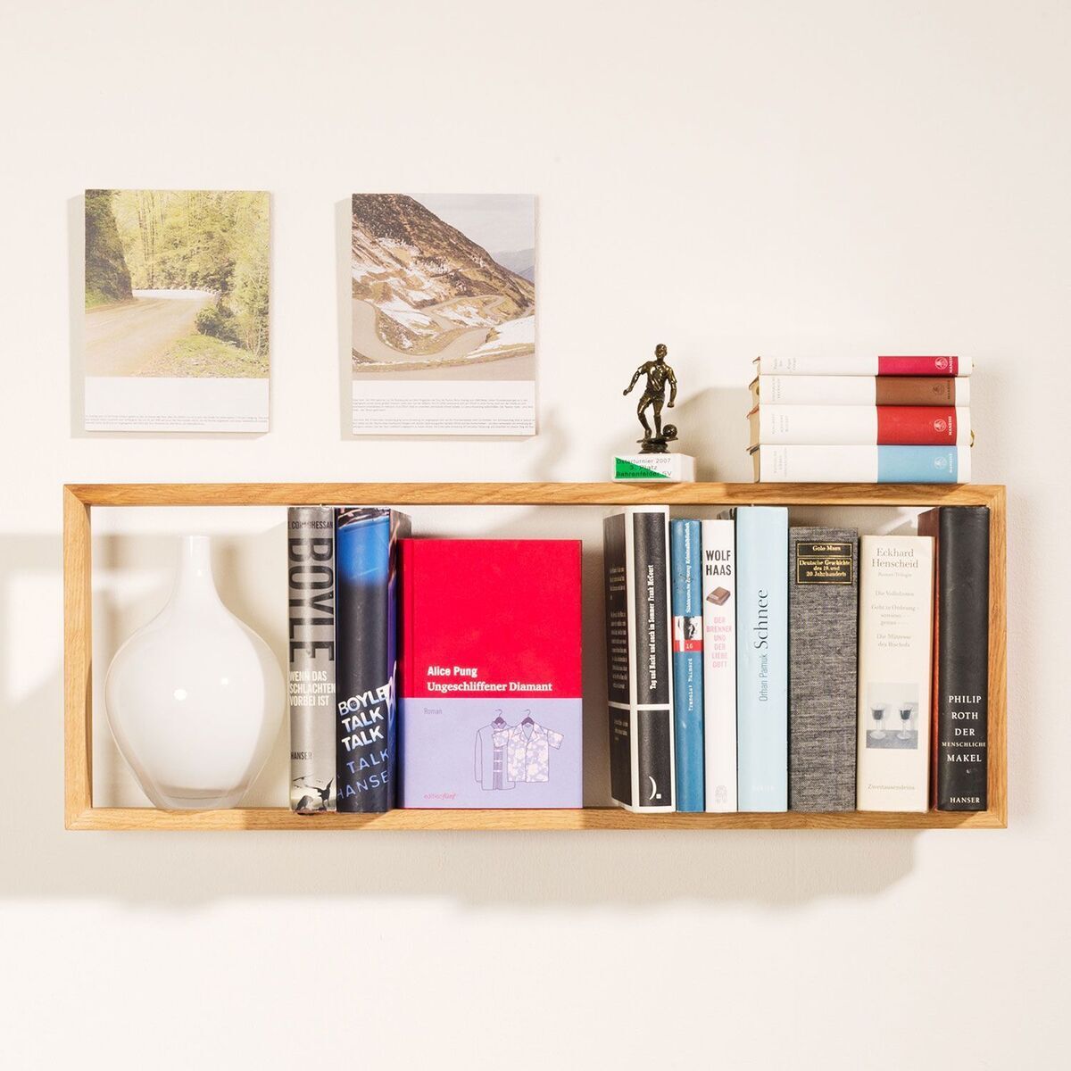 Ein wandmontiertes Regal aus hellem Holz mit verschiedenen Büchern, einer weißen Vase, zwei kleinen Fotoleinwänden und einer kleinen Skulptur darauf an einer hellen Wand.