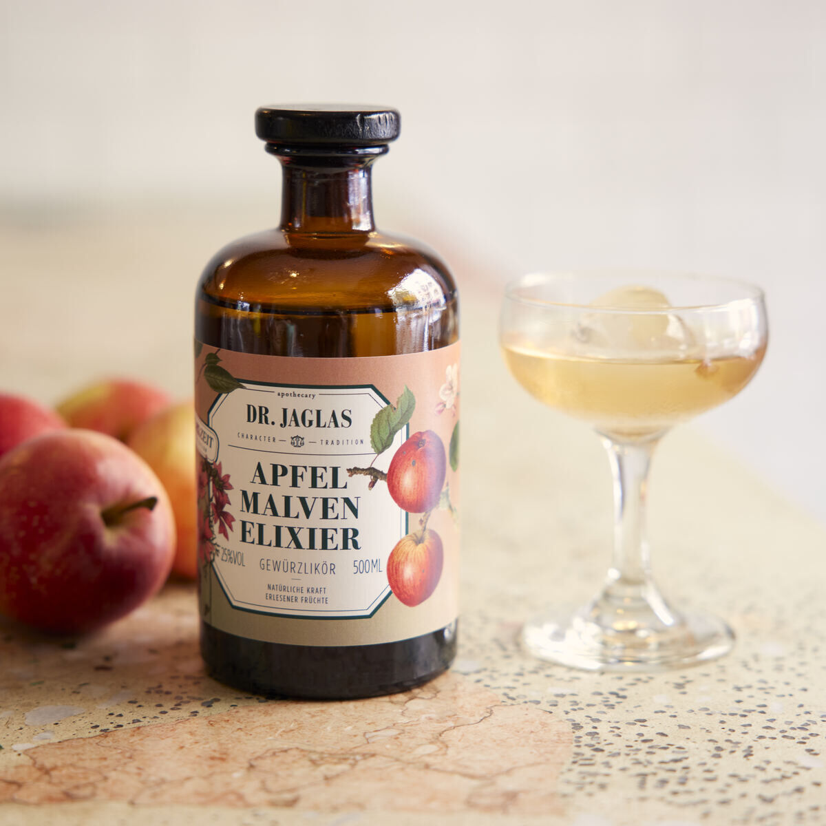 Flasche von Dr. Jaglas Apfel Malven-Elixier Dessertlikör neben einem gefüllten Likörglas und frischen Äpfeln auf einem Tisch.