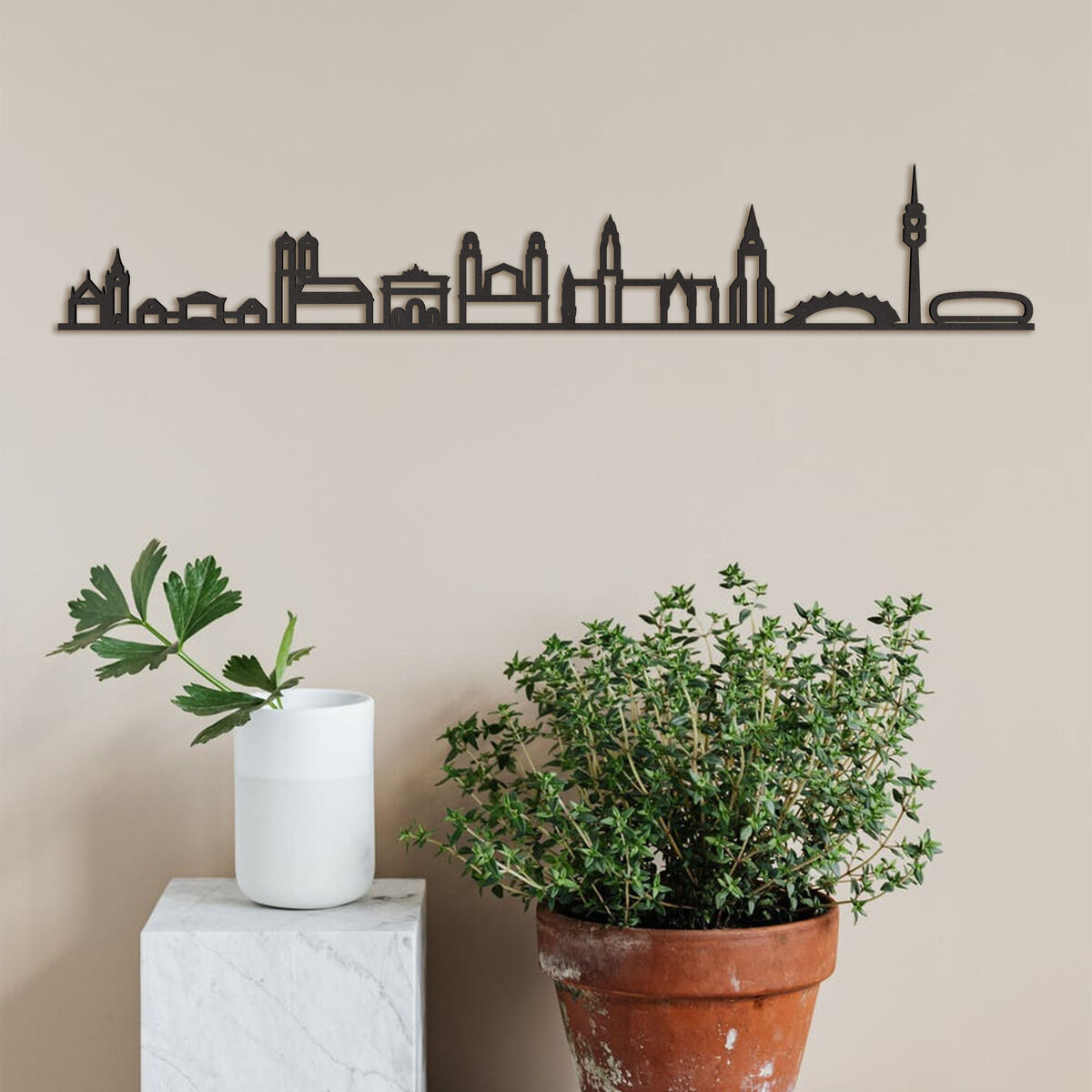 Ein minimalistisches Holzkunstwerk der Skyline von München, das an einer Wand über einer Pflanzen- und Vasenanordnung hängt. Das Design zeigt verschiedene Wahrzeichen der Stadt auf einem schlichten, beigen Hintergrund.