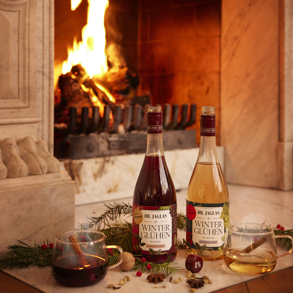 Zwei Flaschen Dr. Jaglas Winterglühen Rot +Weiß alkoholfreier Winzerglühwein stehen vor einem brennenden Kamin aus Marmor. Daneben stehen zwei gefüllte Gläser und winterliche Dekoration wie Tannenzweige und Nüsse auf einem Holztisch. Die Szene strahlt Gemütlichkeit aus.