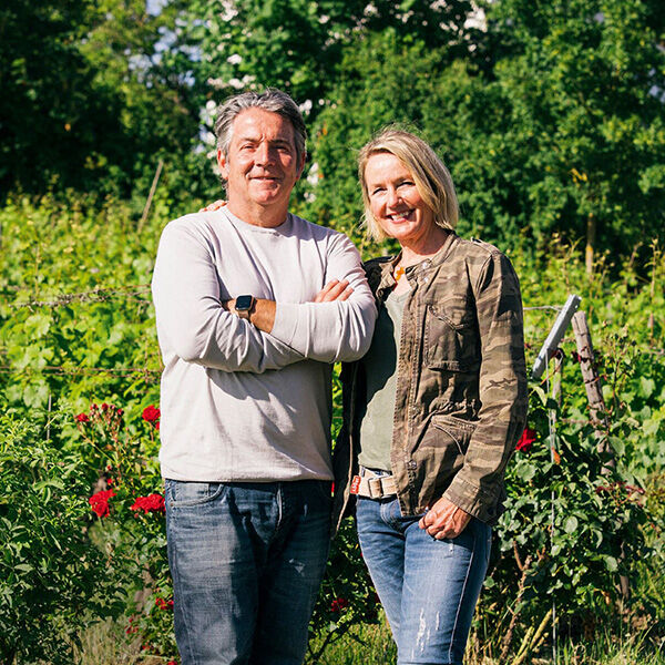 Ein Pärchen steht in einem Weinberg und lächelt in die Kamera. Der Mann trägt ein weißes Langarmshirt und Jeans, die Frau trägt eine grüne Jacke und Jeans. Im Hintergrund sind grüne Weinreben und rote Blumen sichtbar.