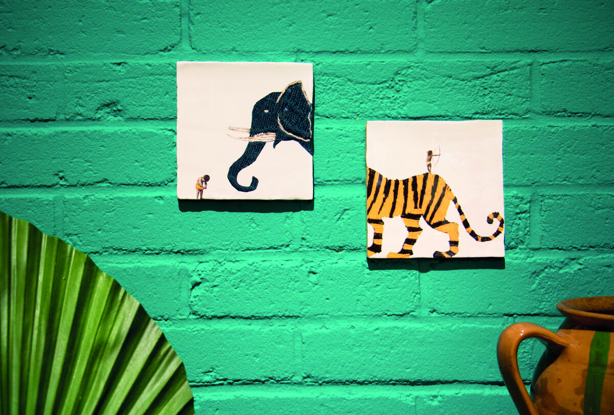 Zwei quadratische Keramikfliesen mit illustrierten Tiermotiven, links ein Elefant und rechts ein Tiger, hängen an einer grünen Ziegelwand. Im Vordergrund sind teilweise ein grünes Blatt und eine braune Vase zu sehen.