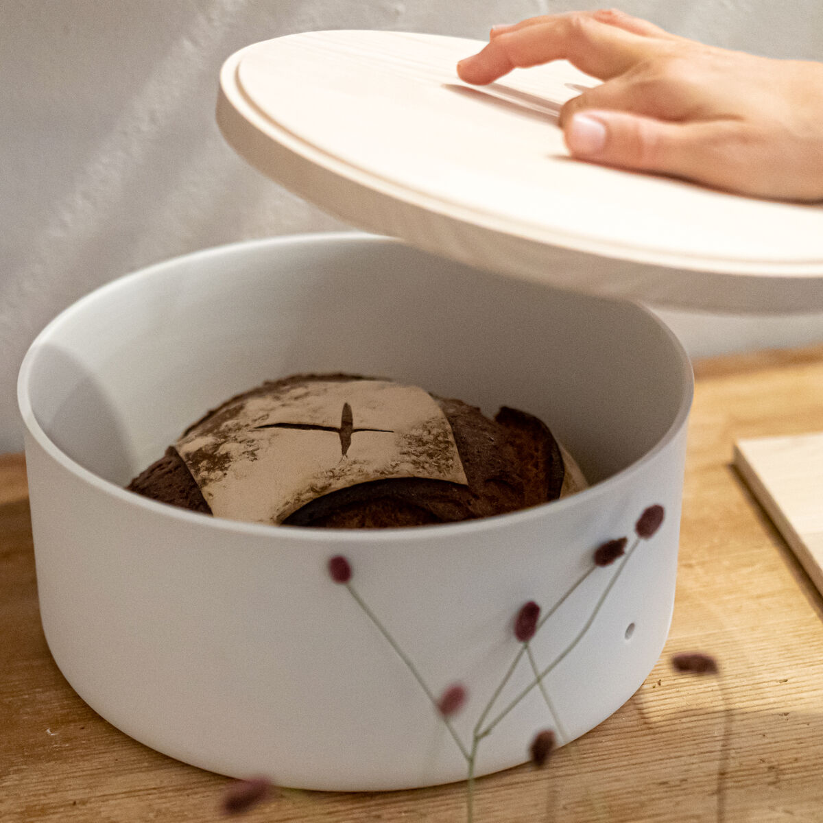 Brottopf "Brot & Brett" von Frau Caze aus weiß mattem Porzellan mit rundem, hellen Eschenholzdeckel, gefüllt mit rustikalem Brot, steht auf einem Holzbrett. Eine Hand hebt den Deckel leicht an. Im Vordergrund einige getrocknete Blüten, dahinter minimalistischer Hintergrund.