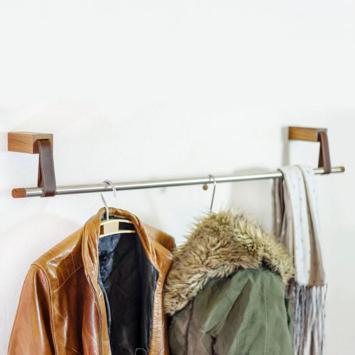 Die minimalistische Garderobe »Ansa« von designimdorf ist an einer weißen Wand montiert. Sie besteht aus zwei eichenholzfarbenen Wandhalterungen mit braunem Leder und einer Edelstahlstange, an der Leder- und Winterjacken sowie Schals ordentlich aufgehängt sind.