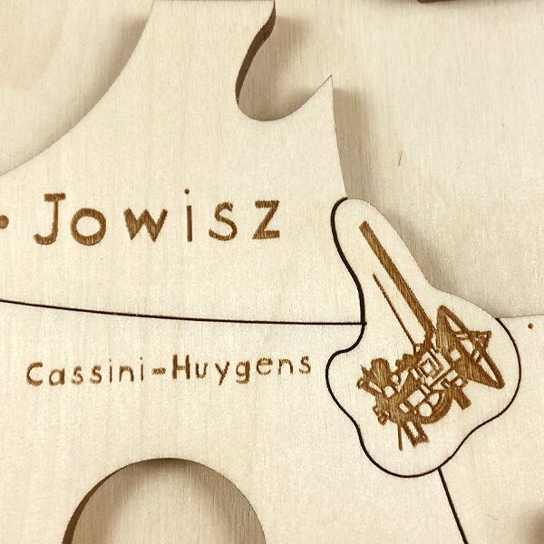 Nahaufnahme eines Holzpuzzles mit dem Thema Weltall. Zu sehen sind beschriftete Puzzleteile mit Aufdrucken von Planeten und einer Raumsonde. Die Farben sind beige, braun und weiß.