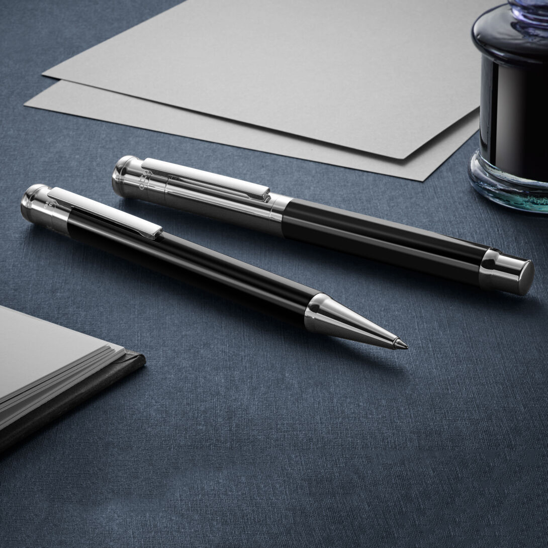 Ein Schreibset mit der Bezeichnung 'Design 04', bestehend aus einem Füllfederhalter und einem Kugelschreiber. Die Schreibgeräte sind in Schwarz und Silber gehalten und liegen auf einem dunklen Schreibtisch. Im Hintergrund sind Blätter und ein Tintenfass zu sehen.