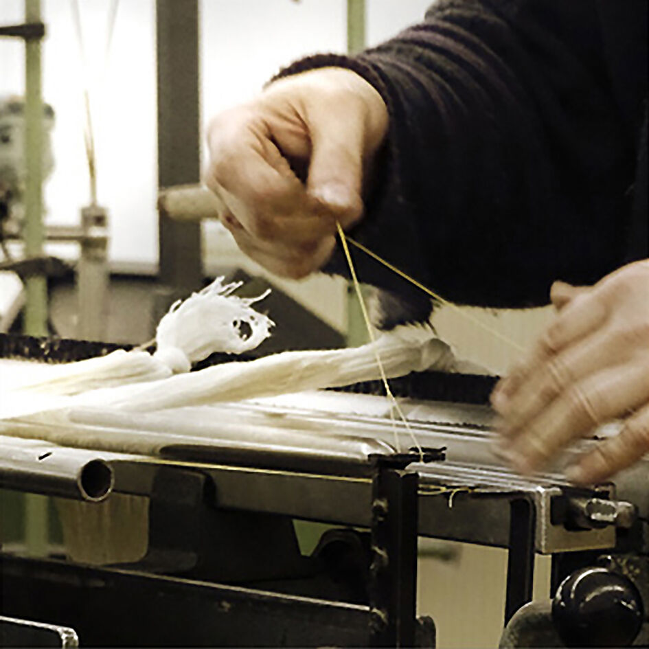 Nahaufnahme einer Person, die an einem gewebten Stoff arbeitet, im Bild ist die Bewegung und Textur der schaffenden Hände erkennbar.