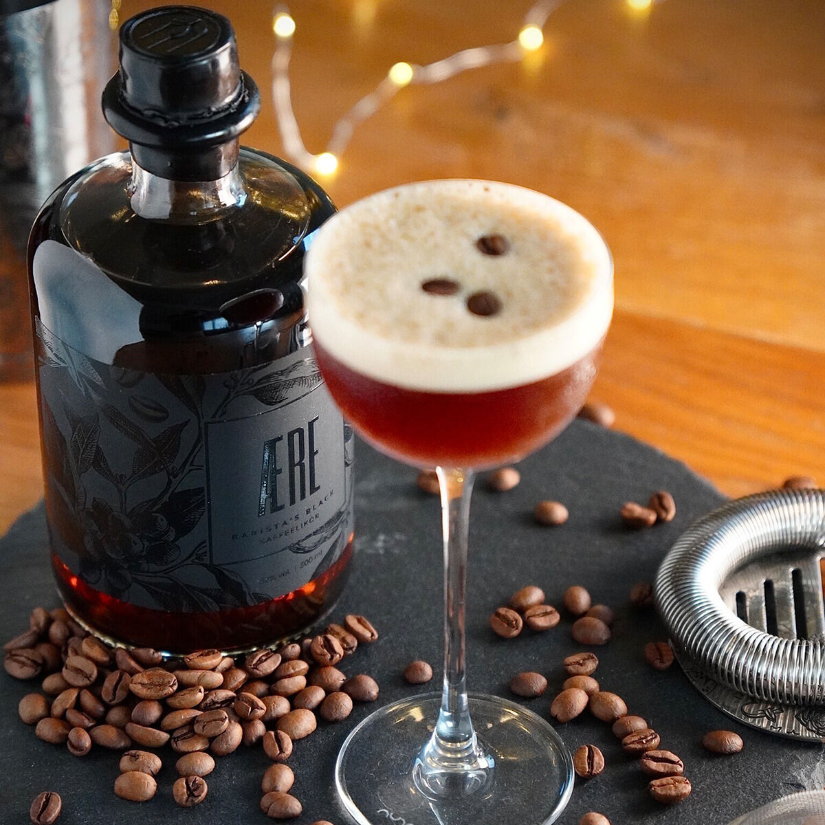 Ein Bild einer Flasche AERE Barista's Black Kaffeelikör, umgeben von Kaffeebohnen. Daneben steht ein Cocktailglas mit einem Kaffeelikörgetränk, das mit drei Kaffeebohnen garniert ist. Im Hintergrund sind unscharfe Lichter zu sehen.