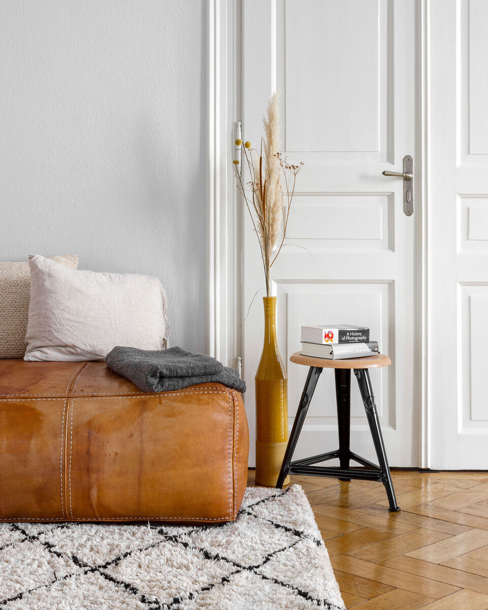 Wohnzimmer mit einem Ledersofa, dekoriert mit Kissen; daneben steht ein kleiner Schemel aus Eiche mit mattschwarzem Rahmen und darauf liegt ein Buch. Im Hintergrund steht eine weiße Tür und eine Vase mit getrockneten Pflanzen. Der Raum hat einen holzfarbenen Parkettboden und einen hellgemusterten Teppich.