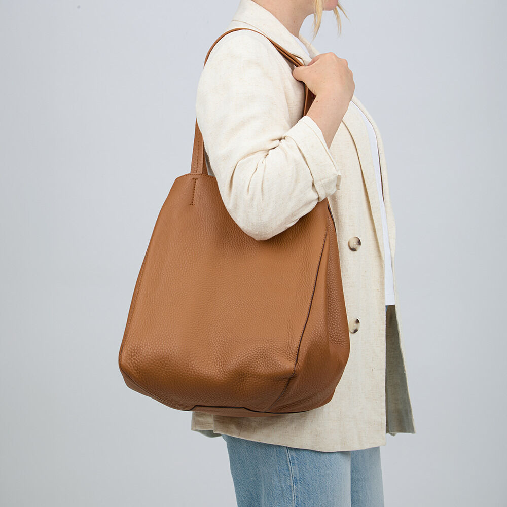 Eine Frau trägt die ZEIT-Tasche »Cep« von Guerer & Guerer in Cognac am Schulterriemen. Die Tasche besteht aus hochwertigem, leicht genarbtem Nappaleder und ist schlicht gestaltet. Der Hintergrund ist neutral grau, die Kleidung der Frau hell.