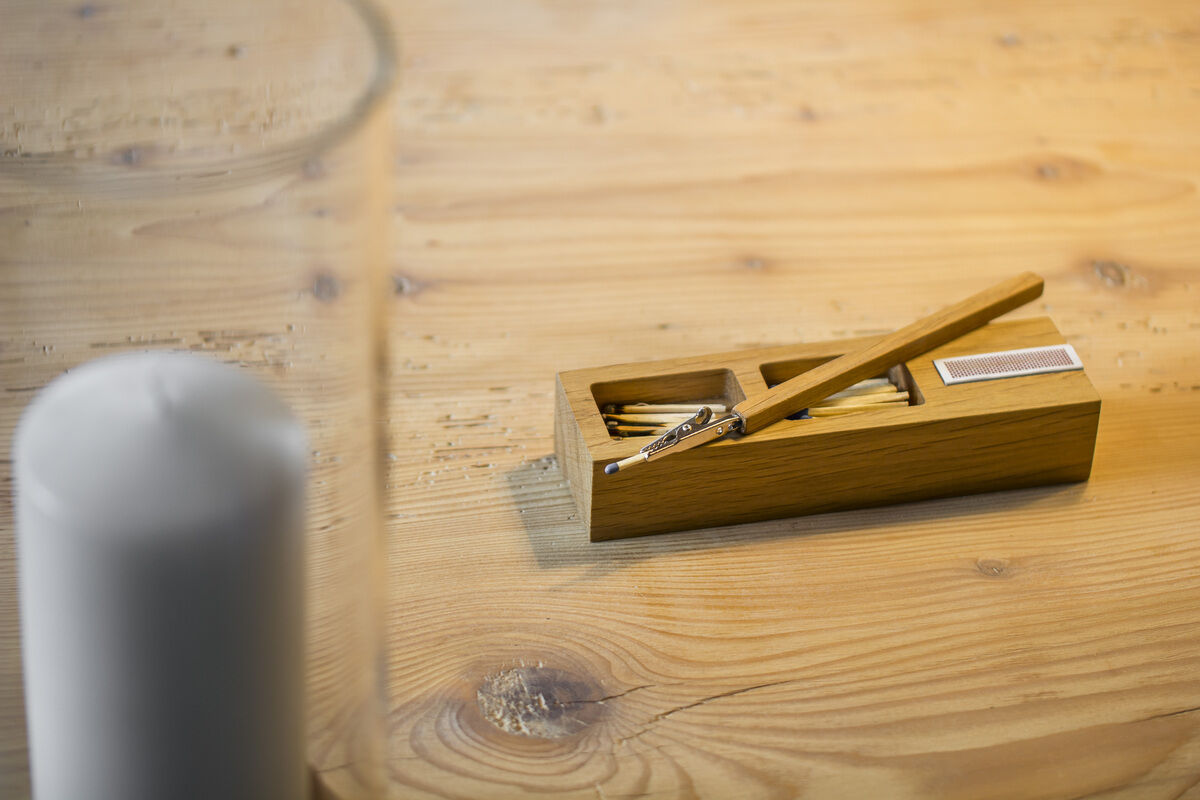 Eine Holzbox aus Eiche, die Streichhölzer und einen kleinen Metalleinsatz enthält, steht auf einem hölzernen Tisch. Daneben befindet sich eine weiße Rundkerze.