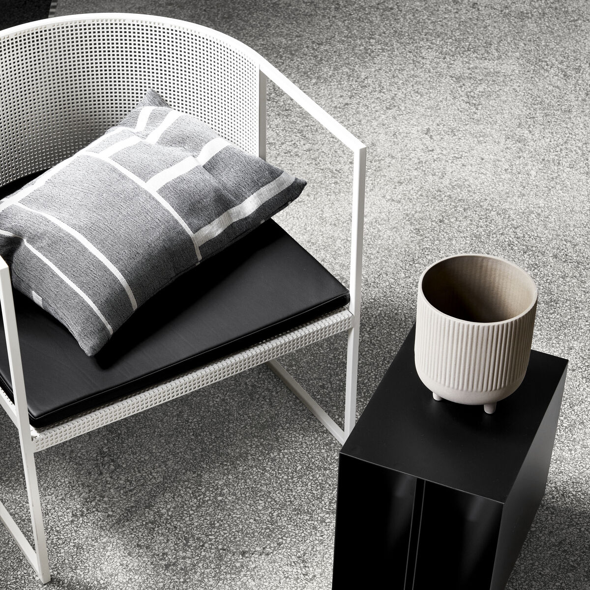Eine stilvolle Wohnzimmer-Szene mit einem modernen weißen Stuhl, auf dem ein grau-weiß gestreiftes Baumwollkissen mit dem Titel »Architecture« liegt. Ein schwarzer moderner Beistelltisch und verschiedene dekorative Gegenstände sind ebenfalls zu sehen.