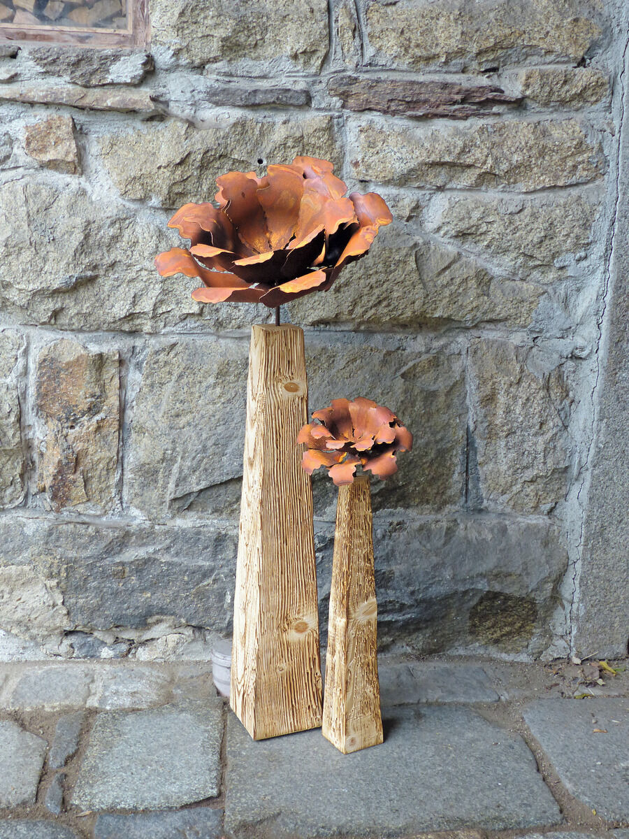 Zwei Gartenstelen mit Kamelienblüten aus Metall vor einer Steinwand. Die Stelen sind aus Holz mit rustikaler Struktur und die Blüten sind in Rostrot und Orange gehalten.