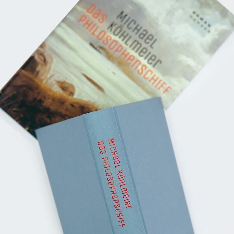 Ein Buchcover des Romans 'Das Philosophenschiff' von Michael Köhlmeier. Der Titel und der Autorname sind in blauer und roter Schrift, vor einem Hintergrund, der eine Landschaft mit Wasser und Himmel zeigt.