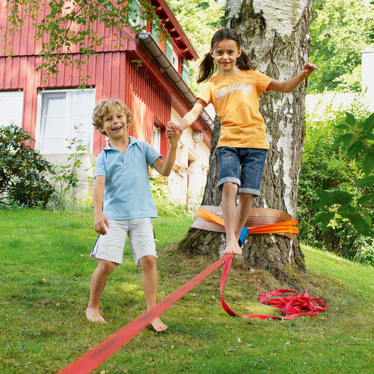 Zwei lachende Kinder, ein Mädchen und ein Junge, balancieren auf einer roten Slackline im Garten. Die Slackline ist an zwei Bäumen befestigt. Im Hintergrund steht ein rotes Haus.