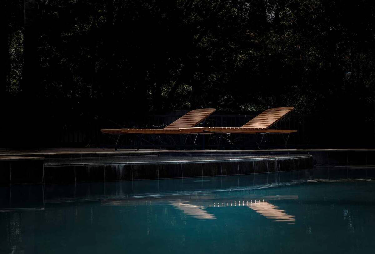 Zwei moderne Liegen der Marke MOLO aus Holz stehen am Rand eines Pools. Die Umgebung ist dunkel mit einigen Bäumen im Hintergrund, die sich im Wasser spiegeln.