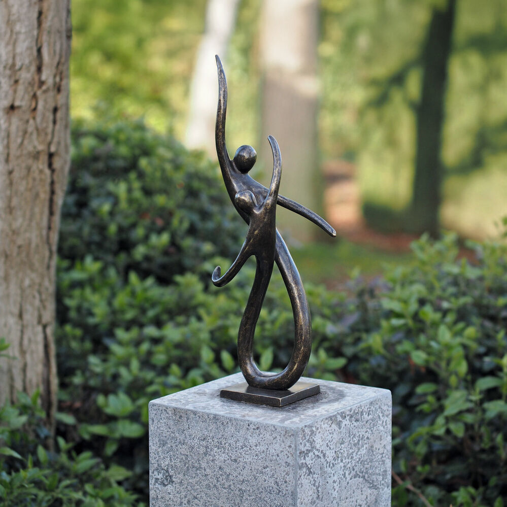 Bronzeskulptur »On the Move« von Ars mundi auf quadratischem Sockel im Garten. Zwei abstrahierte Figuren verschmelzen in einer fließenden Komposition, eine hebt den Arm. Umgeben von üppigem, grünem Pflanzenwuchs. Kunstwerk in moderner Formensprache.