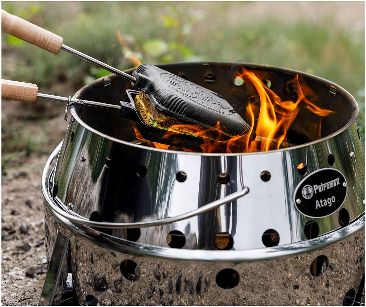 Ein Sandwicheisen mit Holzgriffen wird über einer Feuerstelle auf einem silbernen Grill erhitzt. Flammen lodern um das Eisen, während es ein Sandwich röstet. Im Hintergrund ist eine grüne Grasfläche zu sehen.