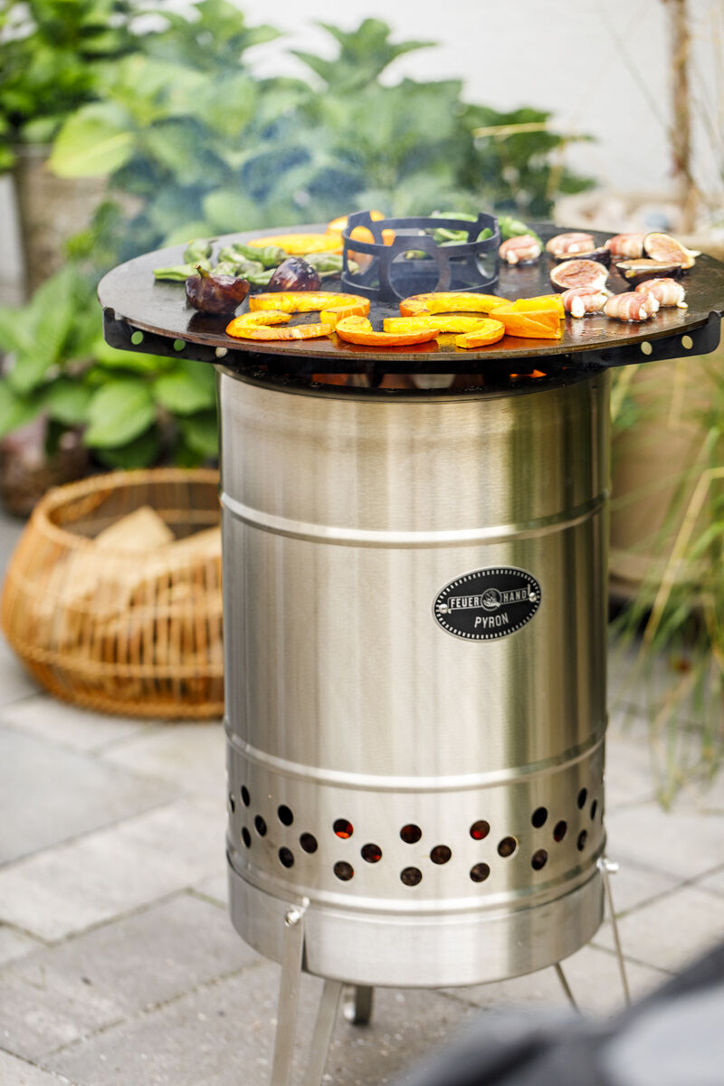 Eine »Pyron« Platte in einem Garten, wo auf der runden Edelstahlplatte verschiedene Gemüse und Fleischstücke gegrillt werden. Im Hintergrund sind Pflanzen und Gartenzubehör zu sehen.