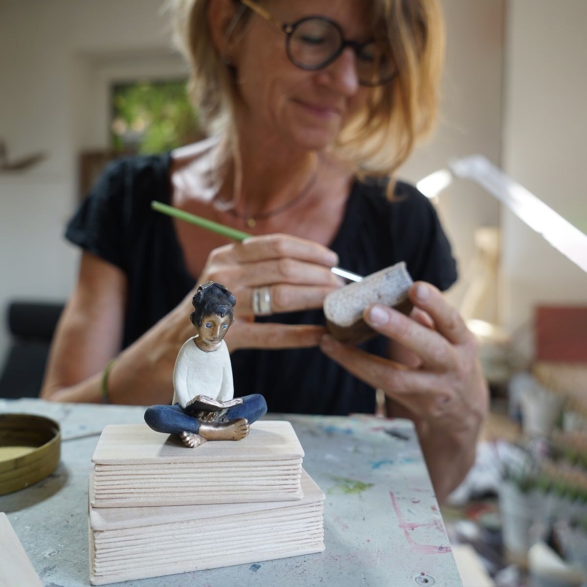 Eine Frau arbeitet an einer Skulptur, die eine lesende Figur zeigt. Die Skulptur steht auf einem Stapel Holzblöcke. Der Name der Künstlerin ist Tamara Suhr.
