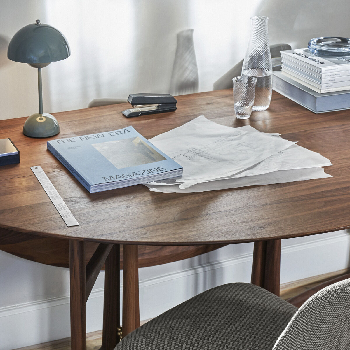 Ein moderner Schreibtisch mit einer Flos Flowerpot VP9 Tischleuchte, Dokumente, Bücher und Dekorationsartikel auf einer Holzoberfläche und ein gepolsterter Stuhl davor.