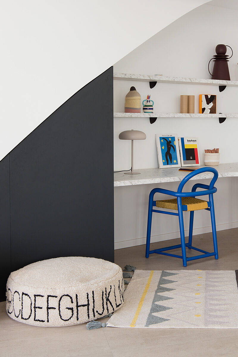 Ein moderner Raum mit einem weißen, mit Buchstaben bedruckten Sitzkissen, einem blauen Stuhl und einem dekorativen Regal im Hintergrund.