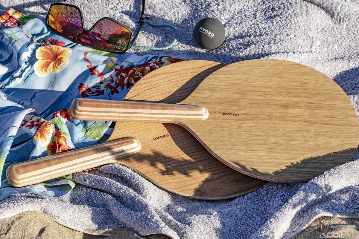 Ein Beachball Set mit zwei Holzschlägern, einem schwarzen Ball, einer Sonnenbrille und einem hellen Strandtuch liegt auf sandigem Boden. Im Hintergrund ist ein blaues Handtuch mit buntem Blumenmuster.