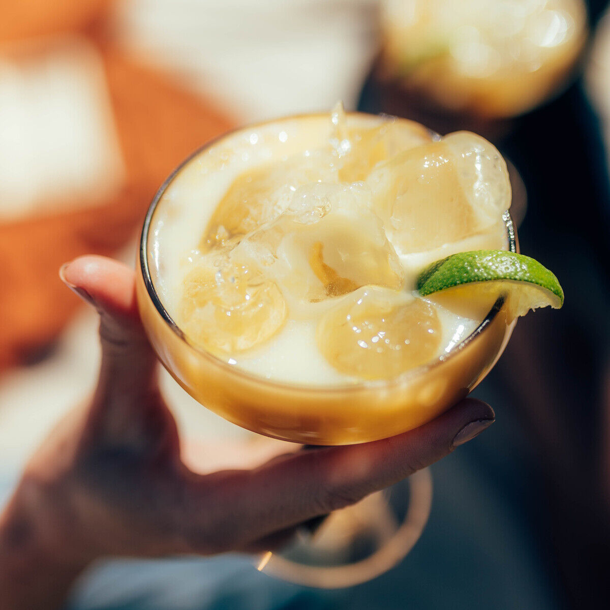 Ein Glas mit cremigem Eierlikör, serviert auf Eiswürfeln und mit Limettenscheibe und Bananenscheiben garniert, wird im Sonnenlicht gehalten. Im Hintergrund ist ein zweites, ähnlich dekoriertes Getränk zu erkennen. Das Bild vermittelt sommerliche Erfrischung und Genuss.