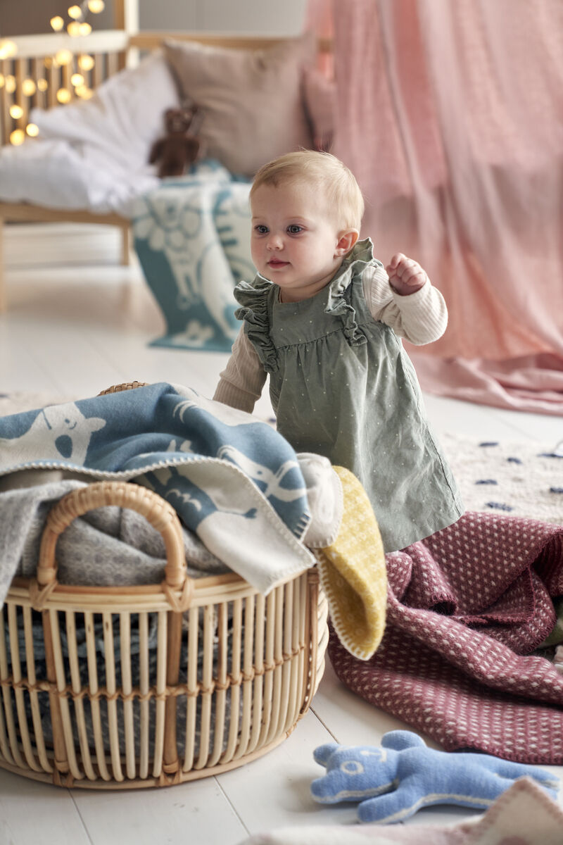 Ein Baby steht neben einem Rattan Korb, der mit mehreren Decken gefüllt ist, in einem hellen, skandinavisch gestalteten Kinderzimmer. Es trägt ein grünliches Kleid, während helle und pastellfarbene Decken und Stofftiere sichtbar sind.
