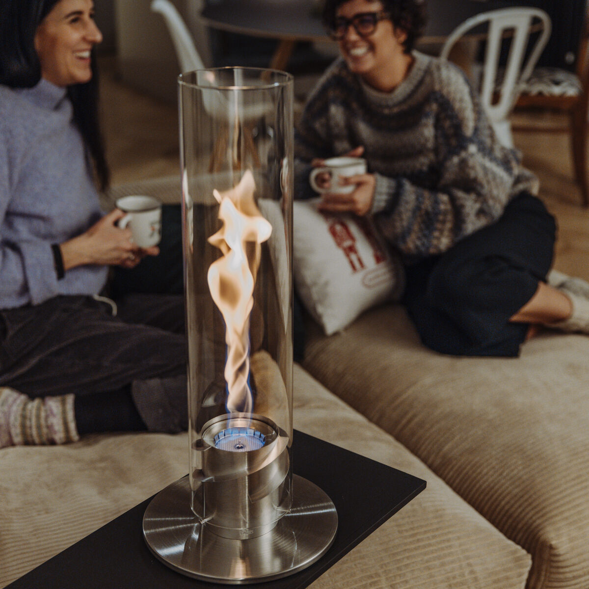 Zwei Frauen sitzen gemütlich in einem Wohnzimmer auf einem Sofa und genießen eine Tasse Kaffee, während ein Tischfeuer »SPIN 1200« in einem modernen, transparenten Metallzylinder sanfte Flammen im Vordergrund erzeugt. Im Hintergrund sind weitere Personen an einer Kücheninsel zu sehen.