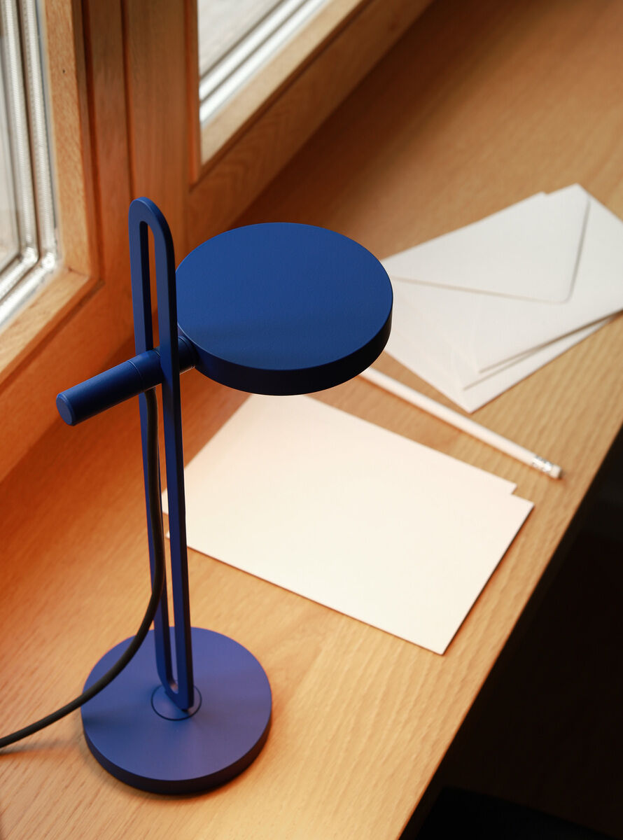 Eine blaue Tischlampe der Marke »Echo« steht auf einem Holzschreibtisch neben weißen Umschlägen und einem Stift. Im Hintergrund sind ein Fenster und Tageslicht zu sehen.
