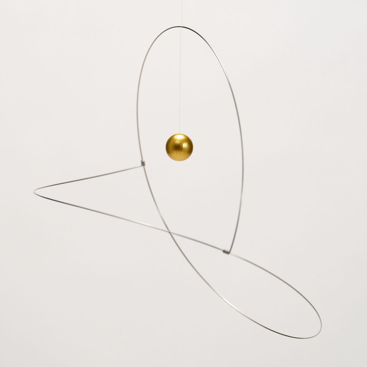 Das Bild zeigt das Mobile "Metamorphosis, Golden Planet" von Flensted Hanging Art. Eine goldene Kugel schwebt im Zentrum zweier eleganter, oval gebogener silberner Metalldrähte vor weißem Hintergrund. Das Design wirkt modern, leicht und minimalistisch.