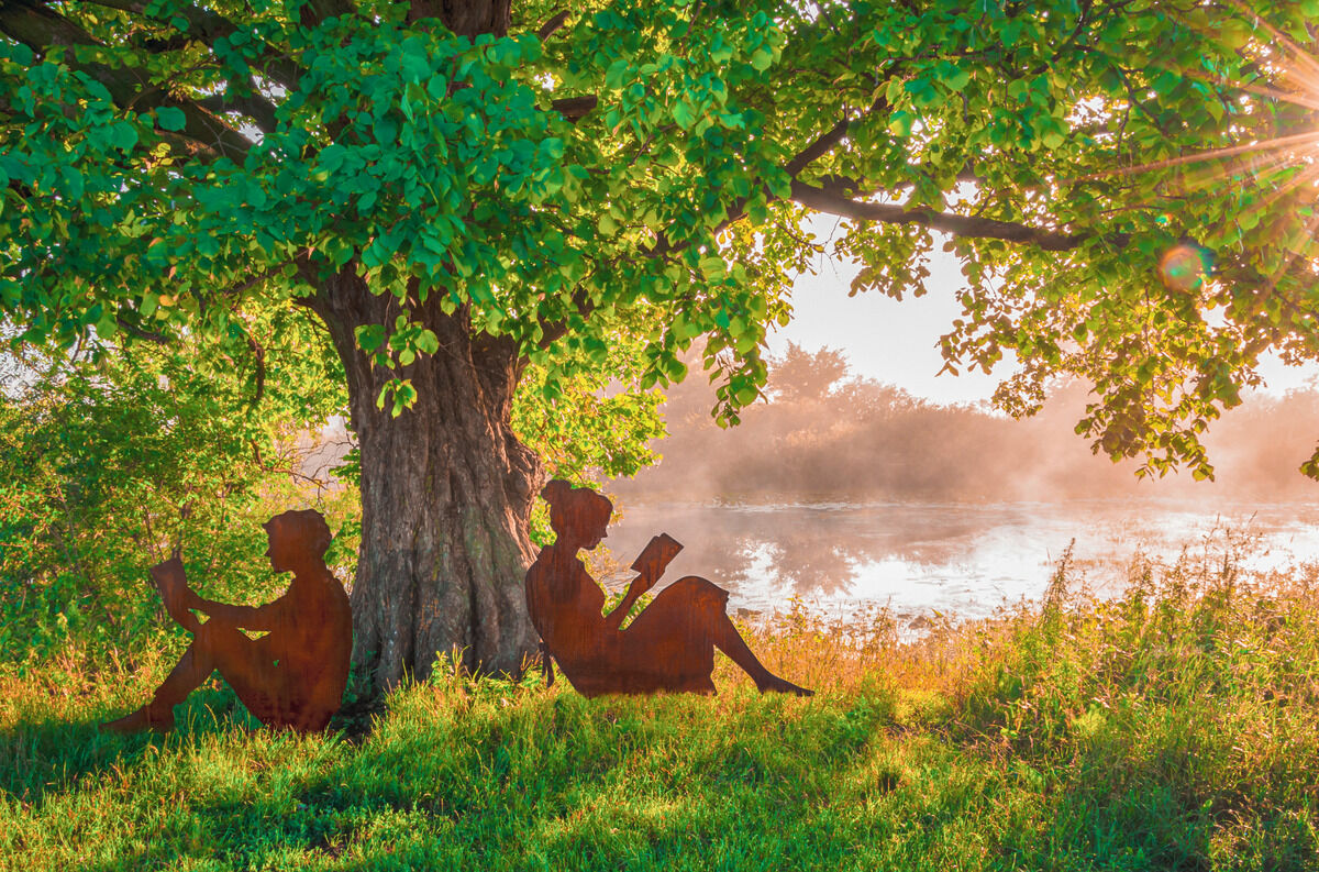 Gartenfigur Silhouette 'Lesende und Lesender' Paar zeigt zwei Silhouetten aus Metall, die unter einem Baum sitzen und lesen, mit einem See im Hintergrund und Sonnenstrahlen durch das Laub.