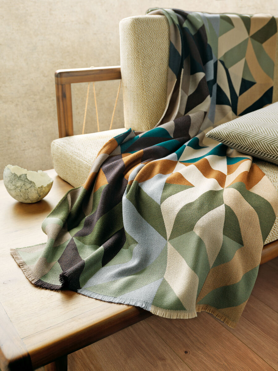 Eine gemusterte Decke »Orlando« liegt auf einem beigefarbenen Sofarücken. Die Decke hat ein geometrisches Muster in verschiedenen Farben wie Grün, Braun, Beige, Schwarz und Blau. Neben dem Sofa steht eine Schale und ein Kissen liegt darauf, was eine gemütliche und stilvolle Atmosphäre im Wohnzimmer schafft.