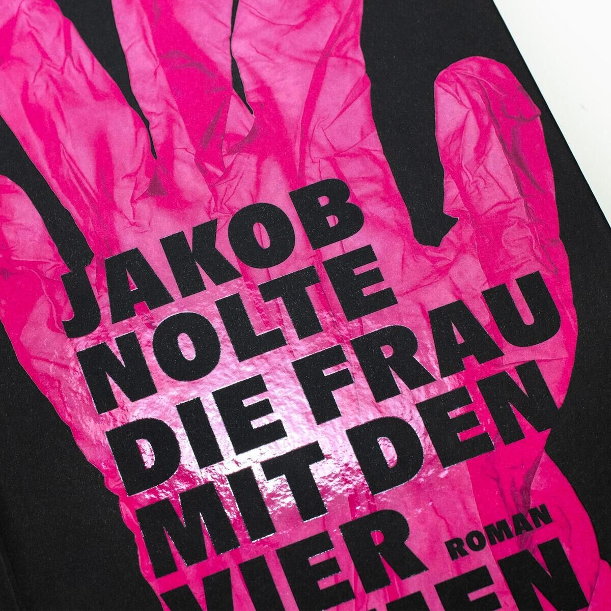 Das Buchcover zeigt eine stilisierte, pinkfarbene Hand mit vier Armen auf einem schwarzen Hintergrund. Der Titel lautet 'Jakob Nolte: Die Frau mit den vier Armen'.