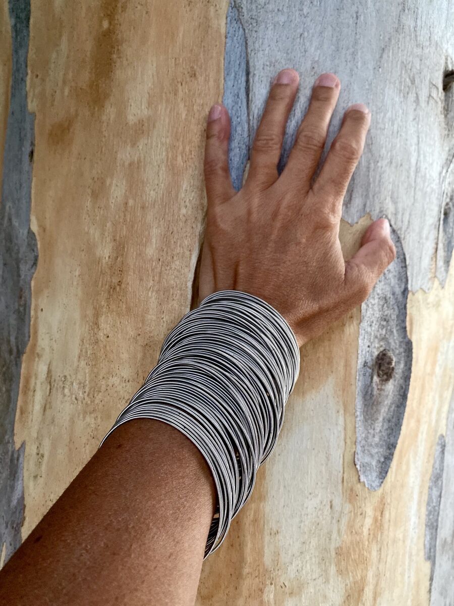 Eine Hand trägt das »Armband 97« aus Edelstahlfedern; das Armband besteht aus mehreren silbernen Drahten, die das Handgelenk umschließen und vor einem Holz Hintergrund präsentiert.