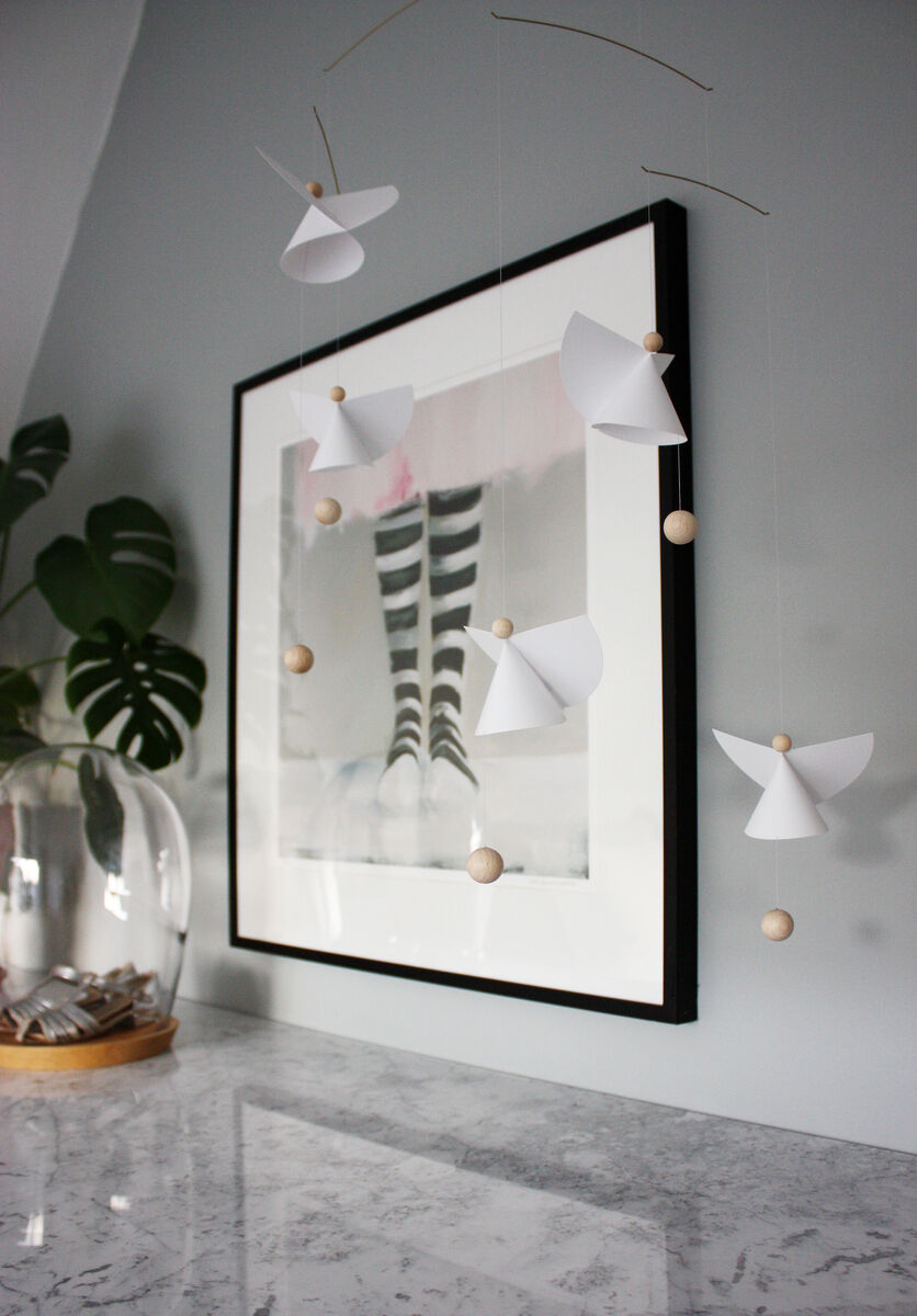 Mobile »Schutzengel 5« bestehend aus weißen Papierengeln und Holzperlen, die an feinen Drähten aufgehängt sind. Das Mobile hängt vor einem modernen gerahmten Kunstwerk und daneben steht eine grüne Monstera-Pflanze in einer glasverschlossenen Vase auf einer Marmorplatte.