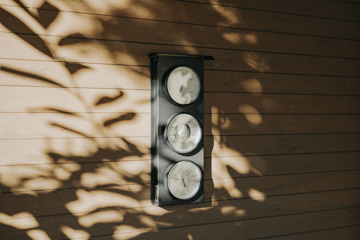 Eine Außenwetterstation der ZEIT-Sonderedition an einer Holzwand montiert. Die Wetterstation enthält drei runde Anzeigen für Barometer, Hygrometer und Thermometer. Sonnenlicht und Schatten von Pflanzen sind auf der Wand zu sehen.